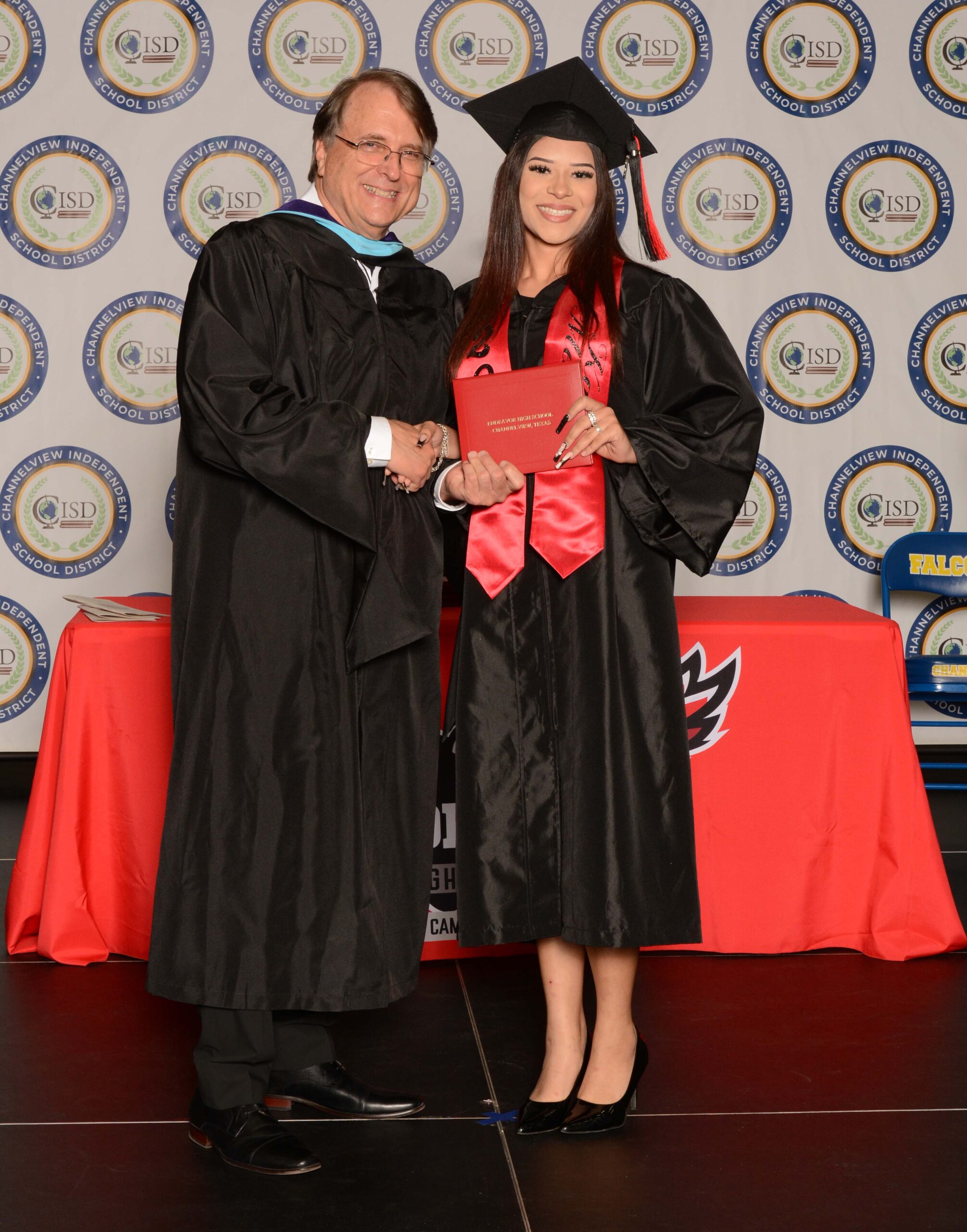 A graduate wearing a black cap and gown holds a diploma folder while standing beside a person in academic regalia on a graduation stage with a patterned backdrop.