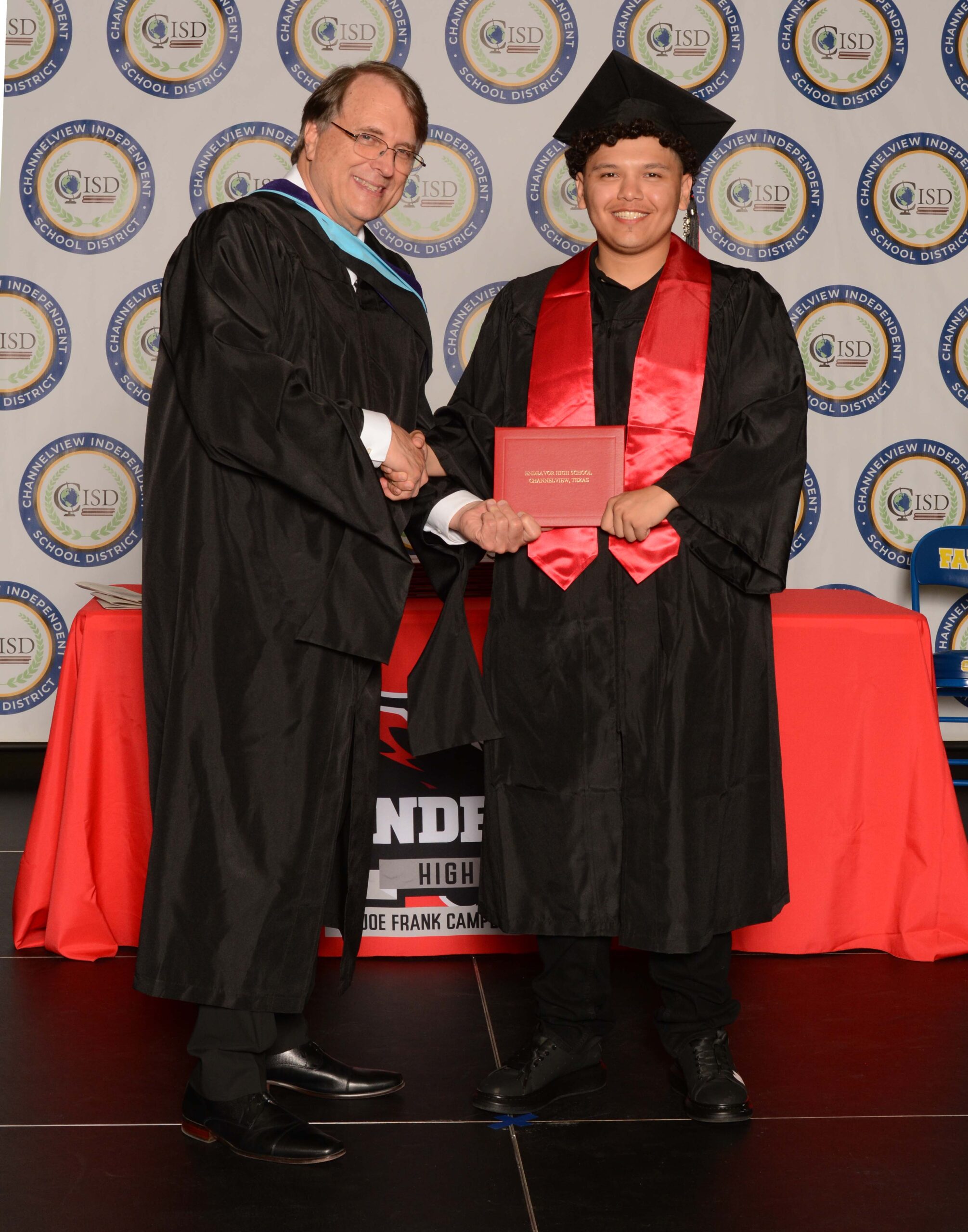 A graduate wearing a black cap and gown holds a diploma folder while standing beside a person in academic regalia on a graduation stage with a patterned backdrop.